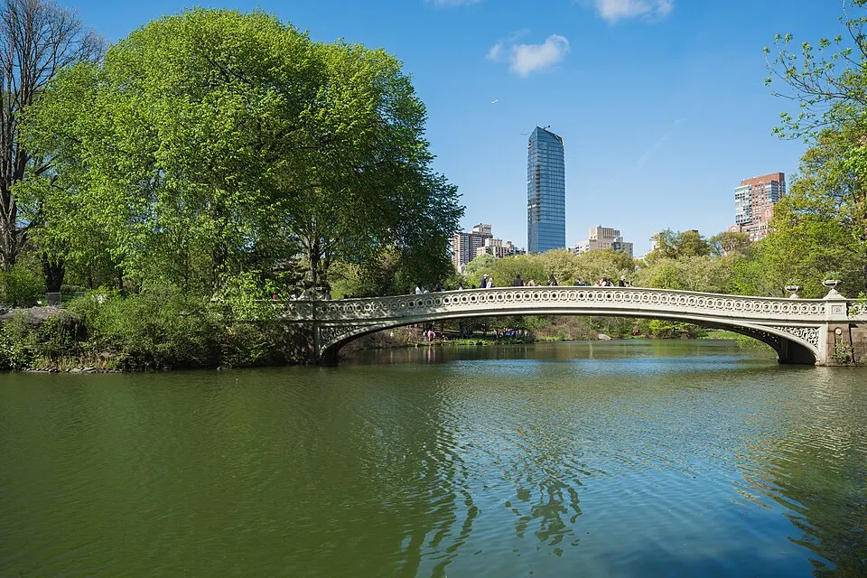 Bow Bridge over the Lake in Central Park, with trees and the Manhattan skyline behind it
